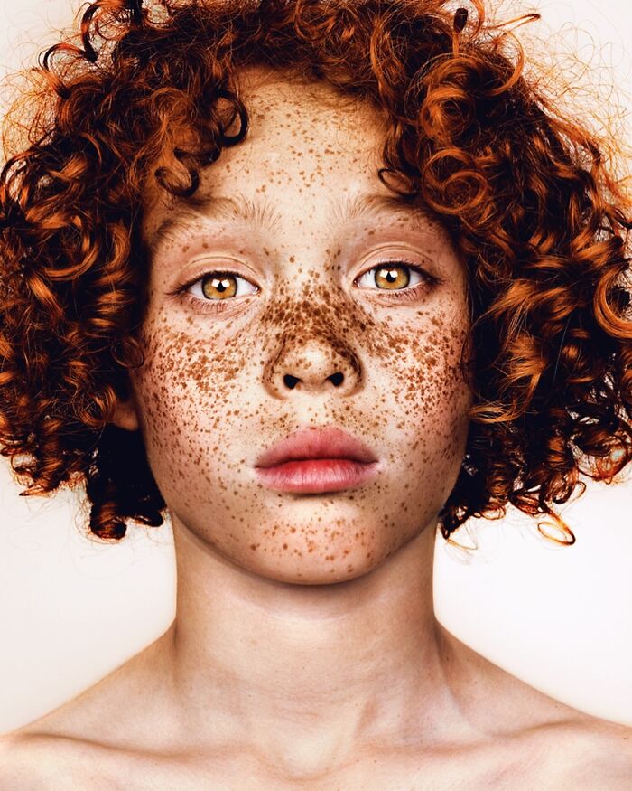 A close-up portrait by Brock Elbank of a child with fiery red curly hair, many freckles, and striking light brown eyes.