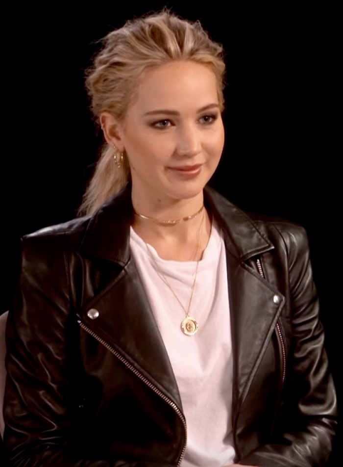 Jennifer Lawrence, with blonde hair tied back, wears a black leather jacket and a white T-shirt. She is smiling faintly, with a black background.