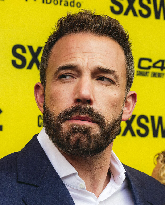Ben Affleck, a famous actor, wears a dark suit and white shirt, looking to the side with a beard, career highlights.