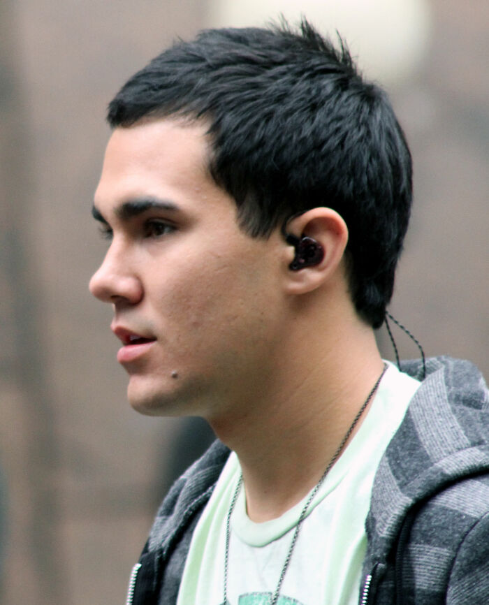 A profile shot of Carlos PenaVega, with an earbud in his ear, highlighting his career highlights and bio.
