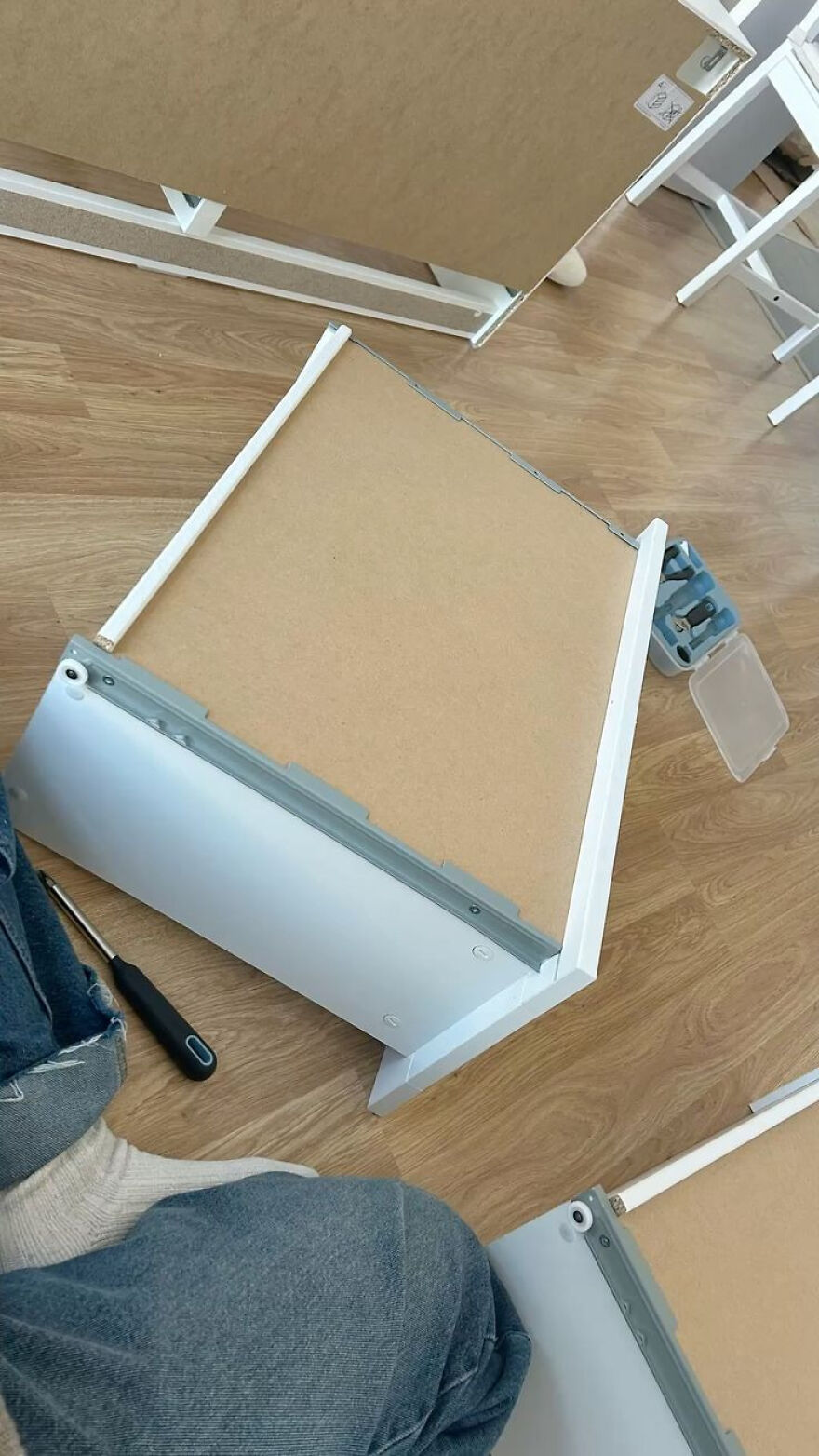 A person sits on a wooden floor next to unassembled furniture pieces and a screwdriver, a weird side hustle.