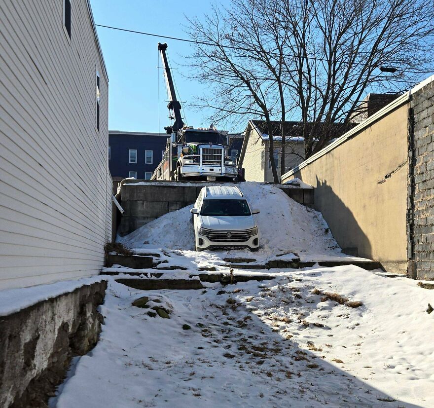 A white SUV stuck on a snowbank, with a large tow truck on top of the hill to help, a funny fail.