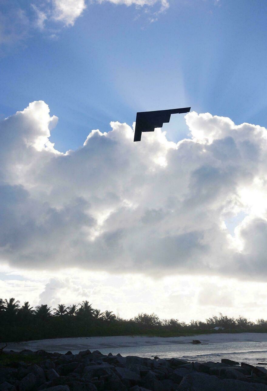Strange thing happening on Earth: A dark, angular object flying above a beach with sun rays behind clouds.