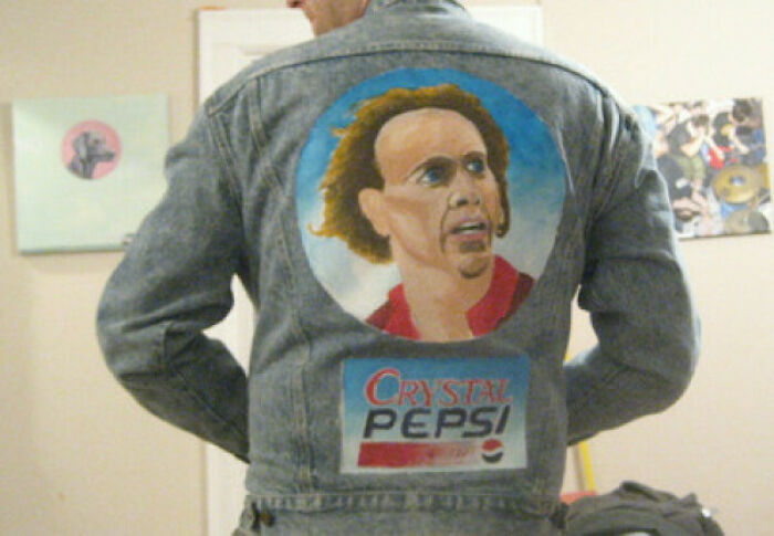 A man wearing a denim jacket with a painted portrait of Nicolas Cage and a Crystal Pepsi logo, a hilarious fashion crime.