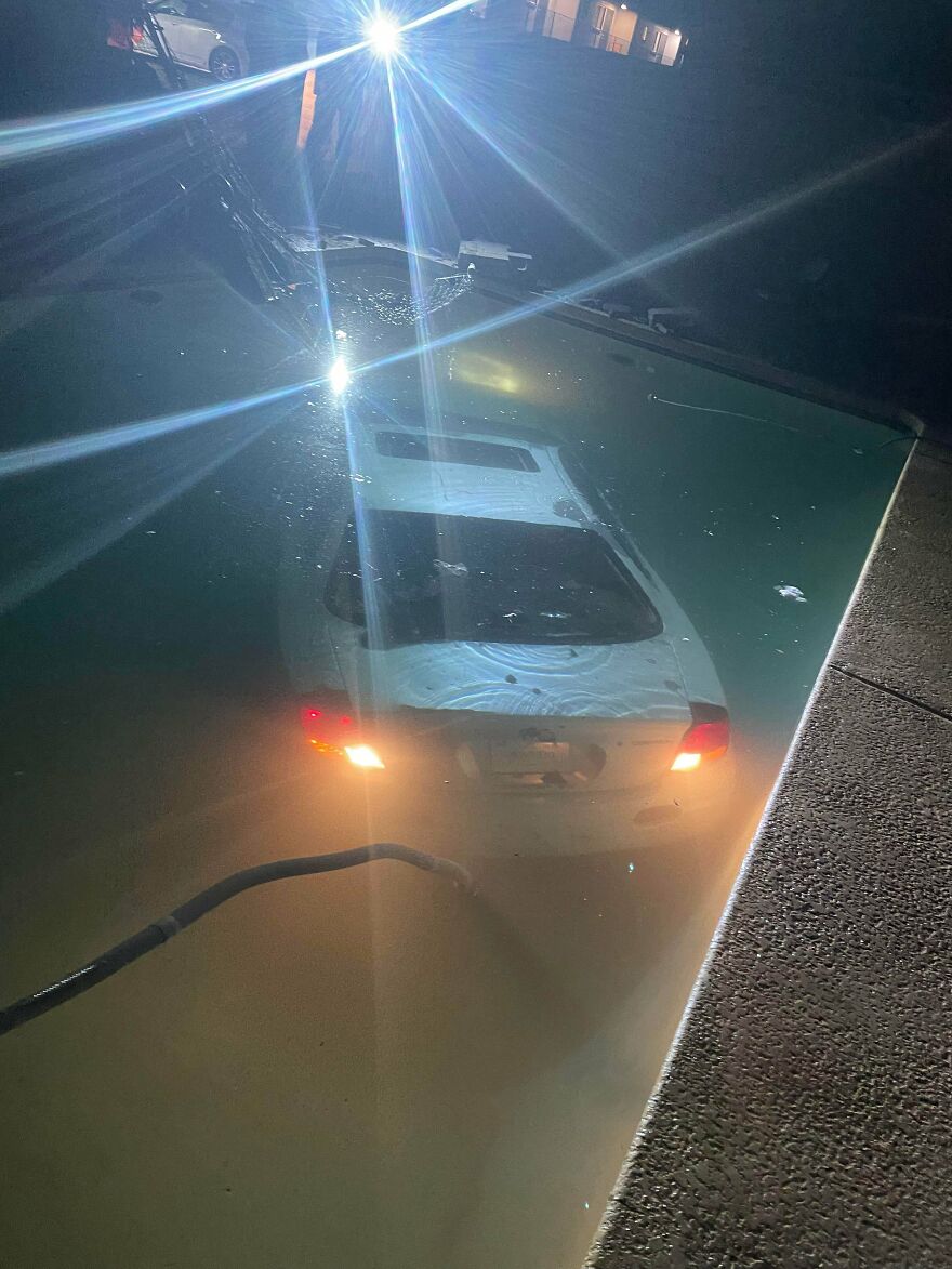 An unfortunate car fail as a white sedan is submerged in a swimming pool at night with its taillights on.