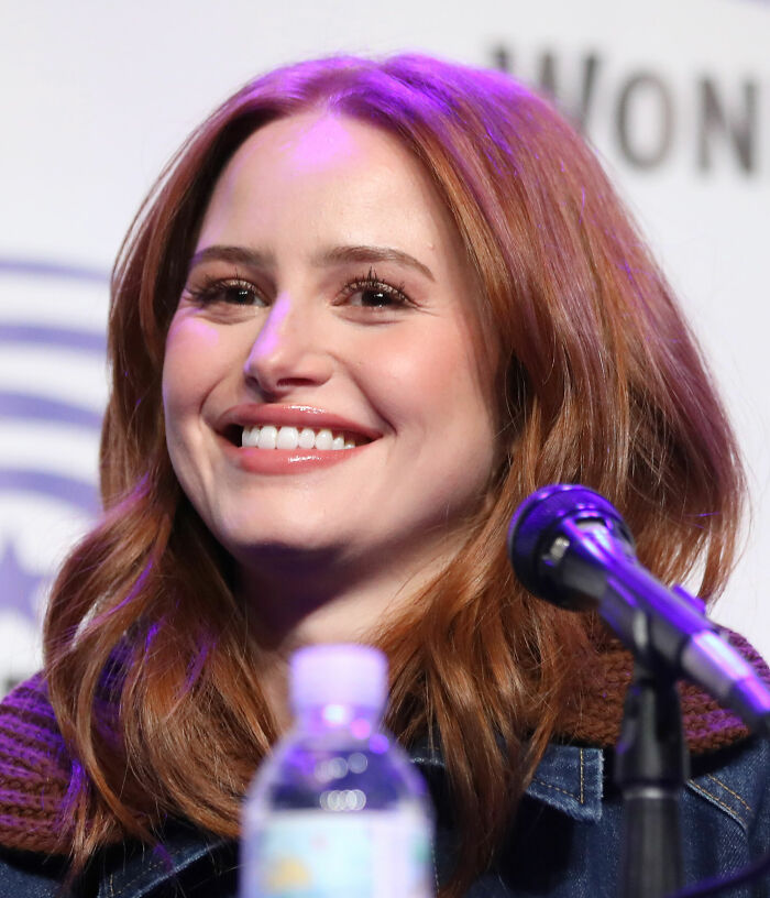 Madelaine Petsch smiling at a conference, showcasing her bright career highlights. The image captures her joyful expression, red hair, and a microphone.