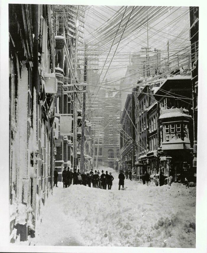 A historic black and white photo showing a snowy city street with numerous power lines overhead.