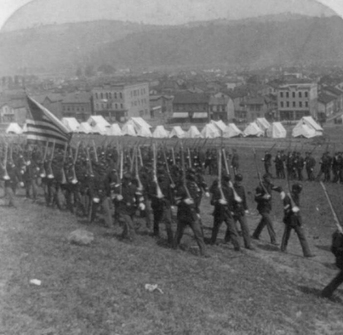 Interesting photo from history of soldiers marching in formation with rifles and an American flag, with tents in the background