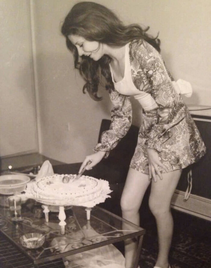 A beautiful photo from history captures a woman in a patterned dress happily cutting a cake on a glass table.