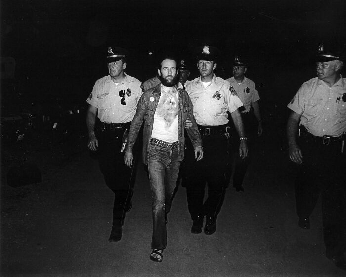 An impactful photo from history shows a bearded man in casual clothes being escorted by several police officers at night.