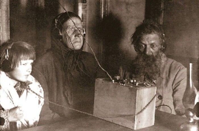A sepia-toned image from history shows a young child, an older woman, and a bearded man wearing headphones, listening intently to a radio.