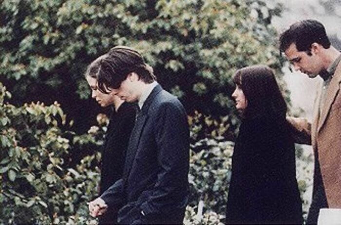 A poignant black and white photo from history features a group of individuals, appearing somber, walking outdoors surrounded by foliage.