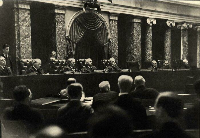 A black and white photo from history shows men seated in a grand hall, possibly a legislative body, with ornate architecture.