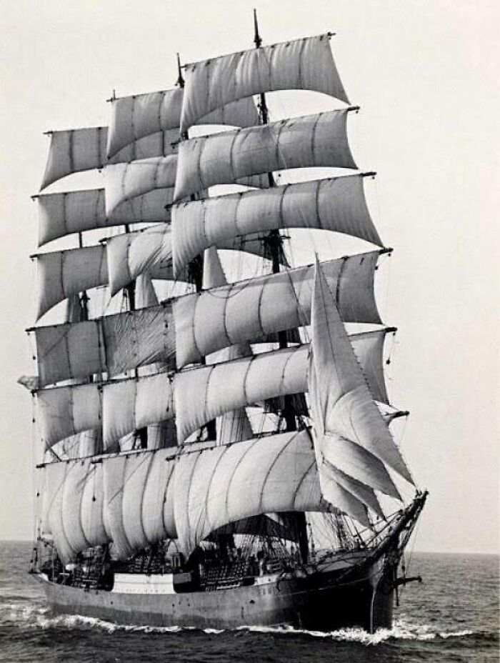 An interesting black and white photo from history of a majestic tall ship with multiple sails unfurled, sailing on the open sea.