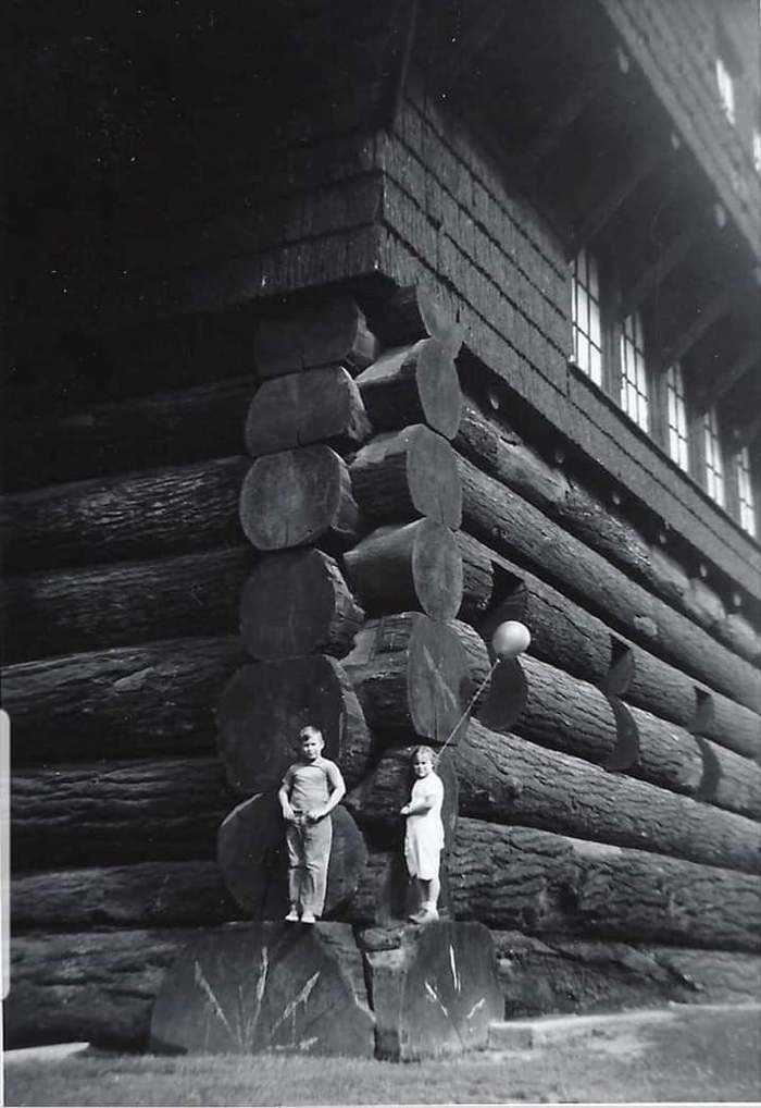 A heartbreaking black and white photo from history showing two children standing on large logs at the corner of a rustic building.