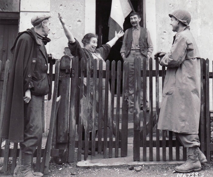 A beautiful black and white photo from history captures a joyful interaction between a soldier and civilians outside a house, with a fence.