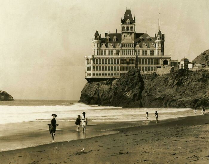 Beautiful photo from history of people on a beach with a grand hotel perched on a cliff overlooking the ocean.