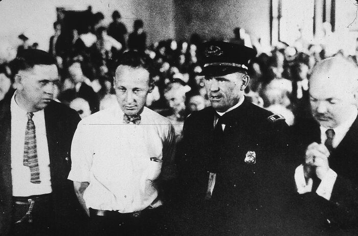 Interesting photo from history of a man in a white shirt and bow tie standing between two other men in a courtroom.