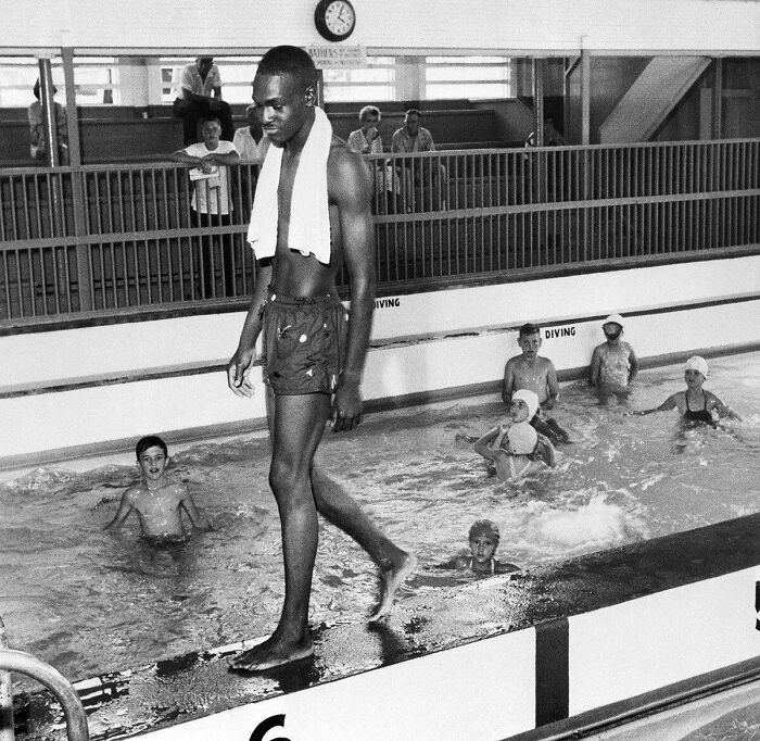 A black man with a towel over his shoulder walks along the edge of a public swimming pool, an interesting moment in history.