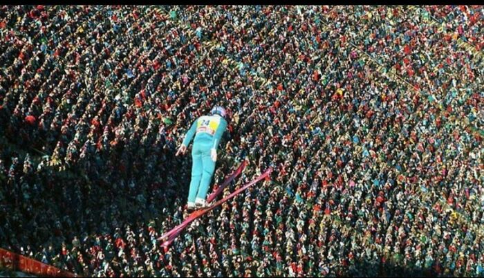 A ski jumper soars over a massive crowd of spectators, captured in a breathtaking and interesting photo from history.