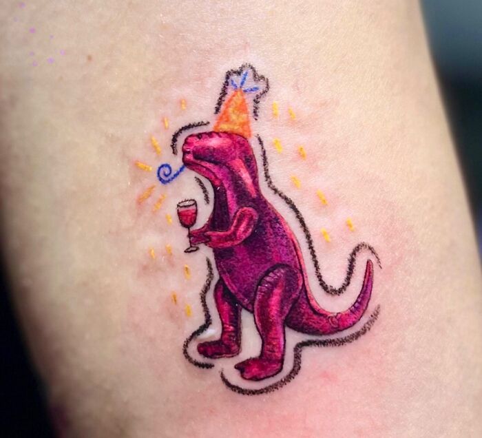 Vibrant crayon drawing-inspired tattoo of a purple dinosaur with a party hat and a drink, reflecting childhood creativity.