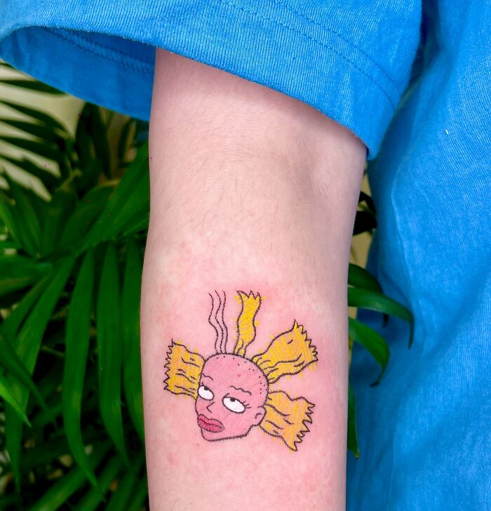 A unique crayon drawing inspired tattoo on an arm, showing a cartoon character's head with pink skin and wild yellow hair, celebrating childhood art.