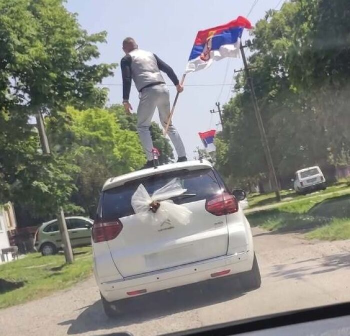 A person standing on the roof of a white car, waving a Serbian flag, showing why people from Eastern Europe are built different.