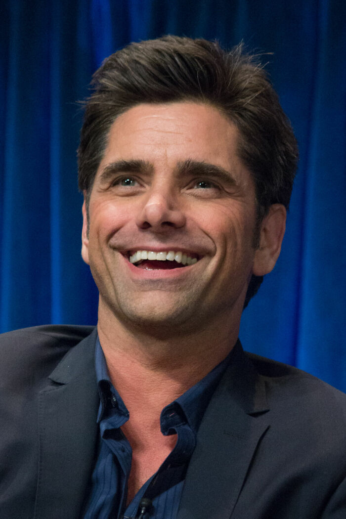 John Stamos smiling, looking happy while wearing a dark suit and open-collar shirt. His career highlights are many.
