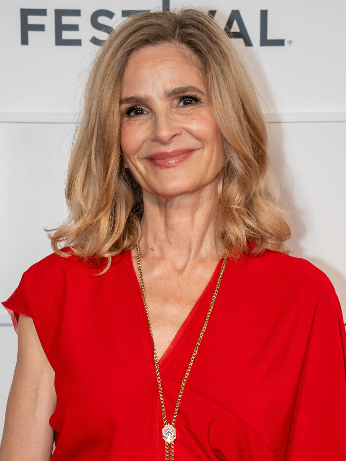 A close-up of Kyra Sedgwick, smiling at the camera, showing her blonde hair and a red top with a golden necklace. Kyra Sedgwick bio and career highlights.