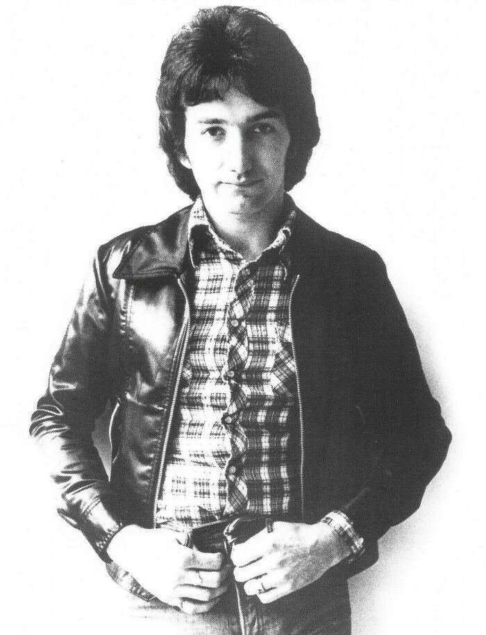 A black and white photo of John Deacon, wearing a plaid shirt and leather jacket, with hands on his belt.