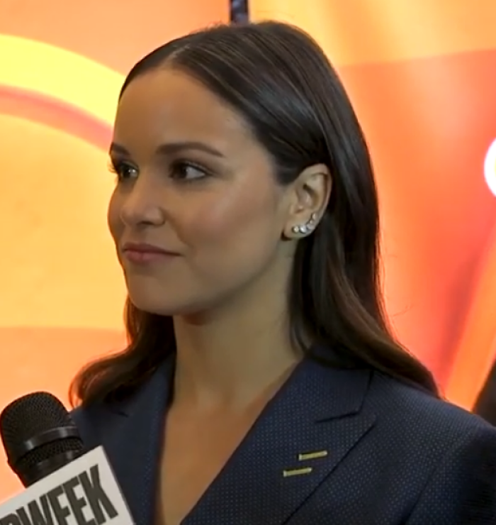 Melissa Fumero with dark hair and a blue suit, being interviewed with a microphone. Her career highlights.