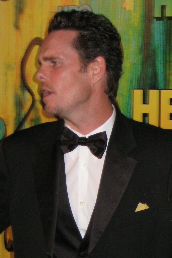 Kevin Dillon in a black tuxedo with a bow tie and a white shirt, looking to the left against a yellow and green background at an event, showcasing his career highlights.