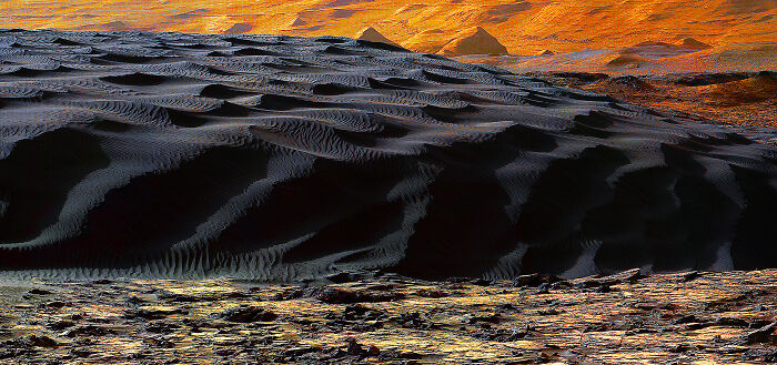 A breathtaking landscape revealing fascinating finds: dark, rippled sand dunes under an orange sky with distant peaks.