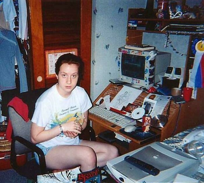 A young person sits at a cluttered desk with an old internet computer, surrounded by 90s era tech.