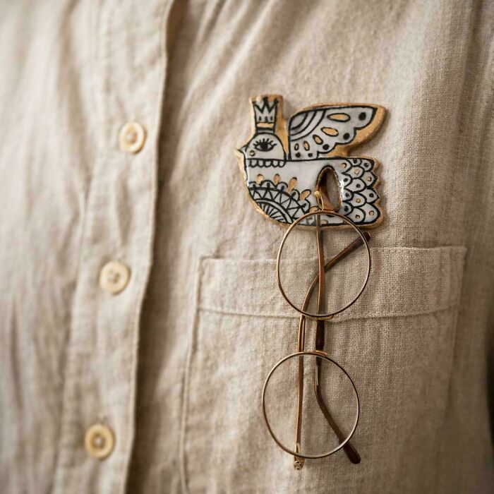 A unique handmade project: a cream-colored bird-shaped brooch with intricate black and gold details on a linen shirt pocket.