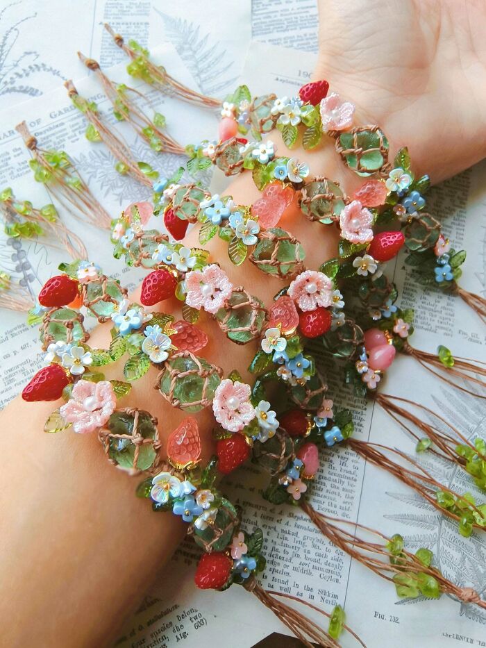 Hands adorned with beautiful handmade projects: bracelets featuring green beads, pink strawberries, and blue flowers.