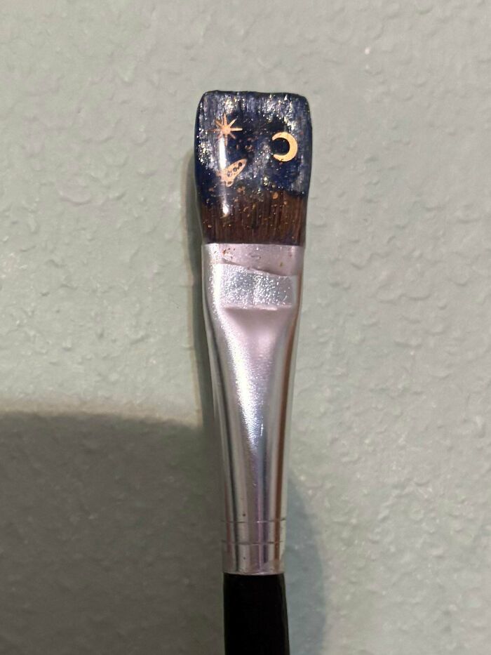A close-up of a handmade project: a paintbrush with its bristles painted to resemble a night sky with a moon and stars.