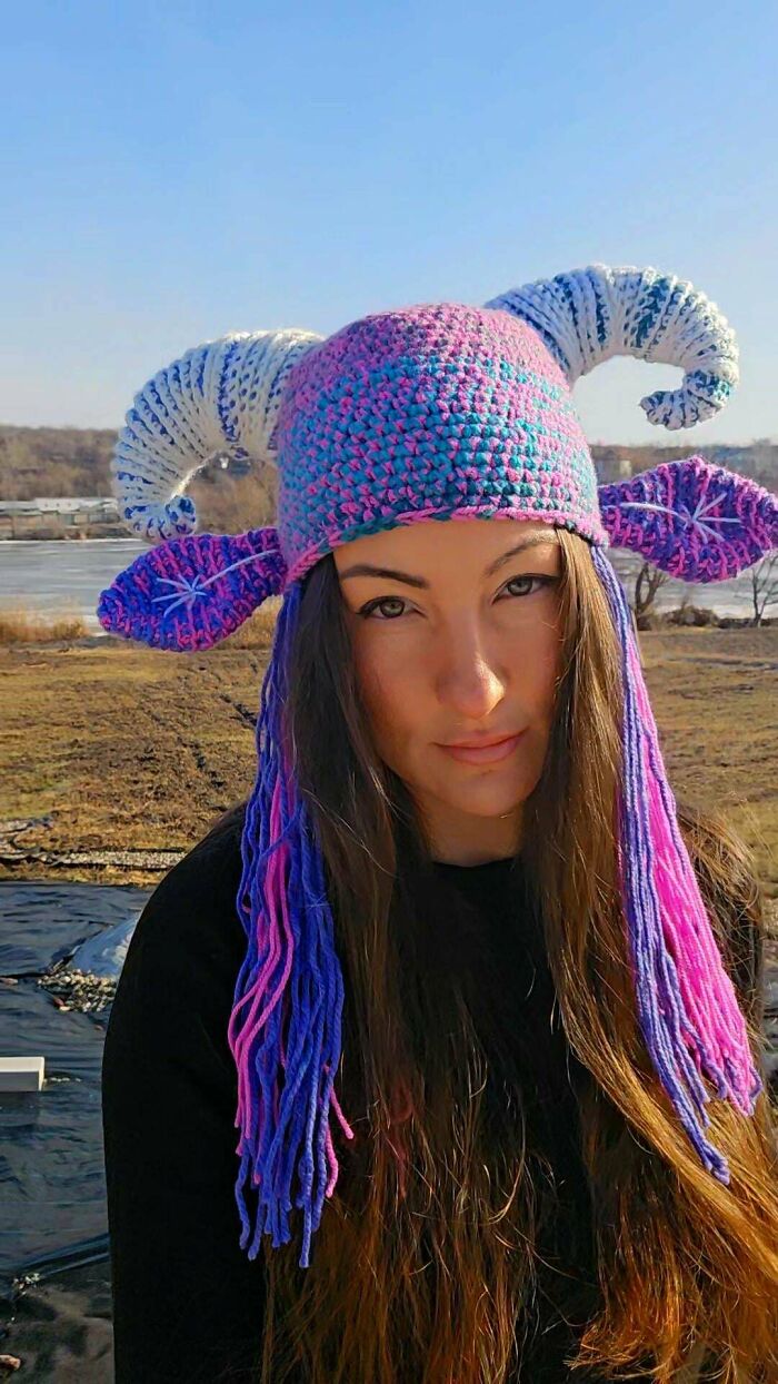 A woman wearing a colorful handmade crochet hat with blue and pink hues, large white horns, and long braided tassels. A unique handmade project.