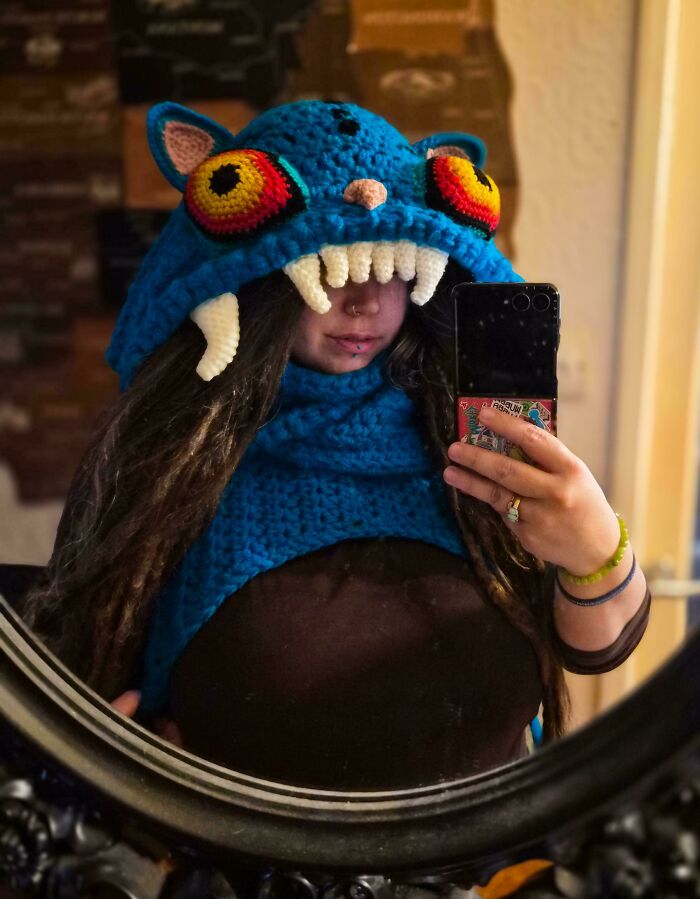 A woman wearing a blue handmade crochet monster hat with large eyes and white fangs. A perfect handmade project.