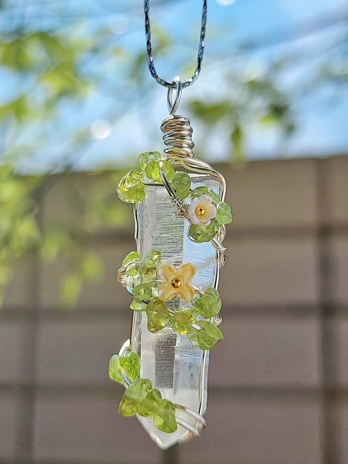 A clear crystal pendant wrapped in wire with green leaf-shaped beads and small white and yellow flowers. A delicate handmade project.