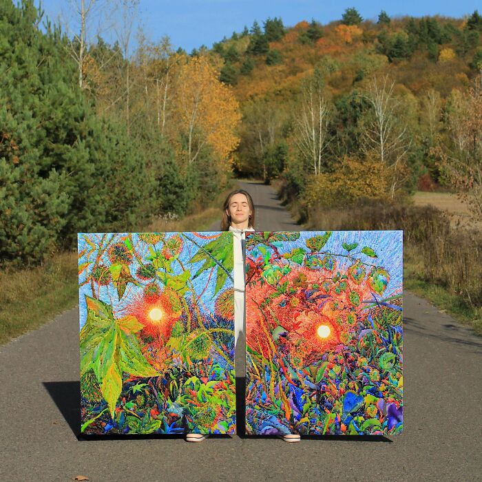 A person holding two large, vibrant handmade projects of nature paintings on a road with a forest backdrop.