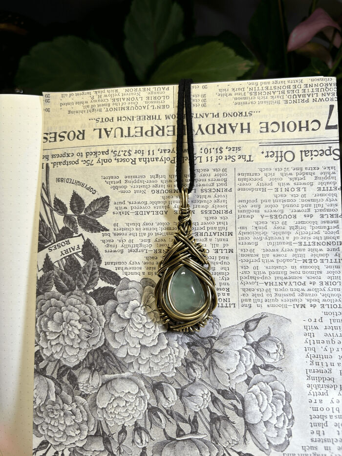 A handmade pendant with a greenish-blue stone wrapped in copper wire, resting on a vintage floral book page.