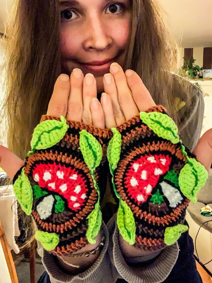 A woman wearing handmade projects: brown fingerless gloves with red mushroom and green leaf designs.