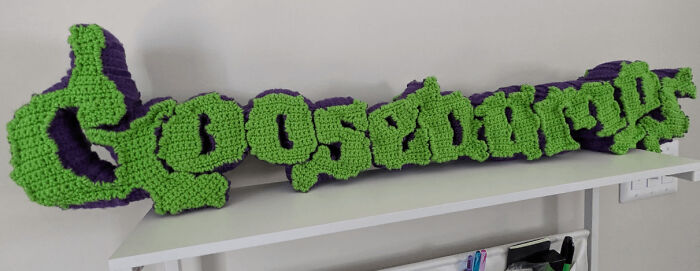 A large, green and purple crocheted Goosebumps logo, a fun handmade project displayed on a shelf.
