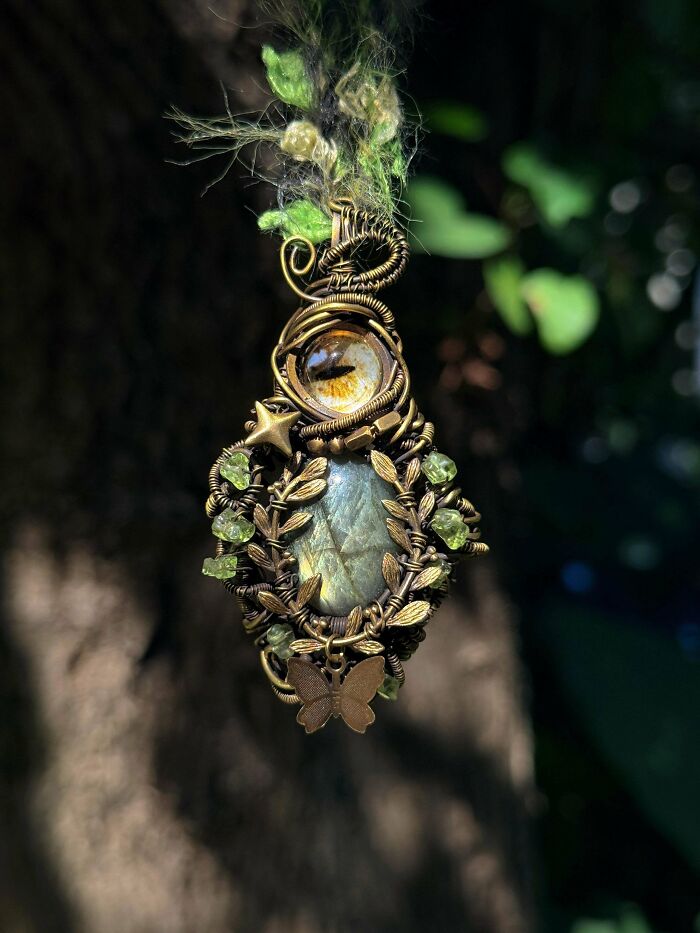 A handmade projects wire-wrapped pendant featuring an eye, an oval stone, and green beads, hanging outdoors.