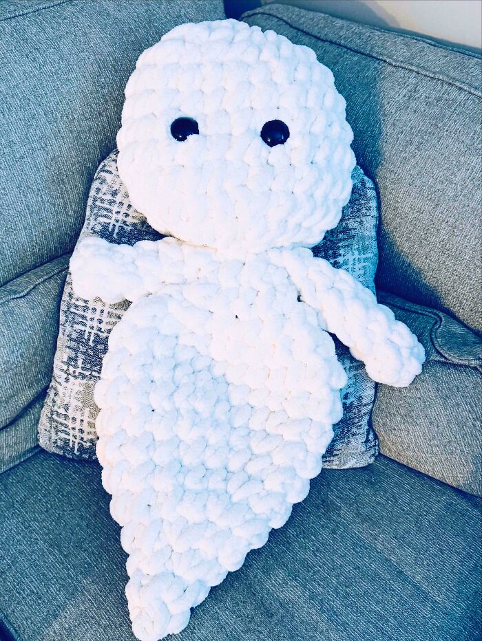 A fluffy white ghost-shaped handmade stuffed animal with black eyes, sitting on a grey couch, a cozy project.