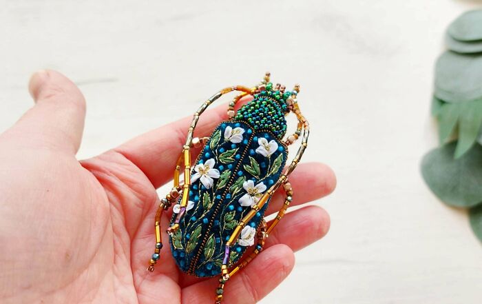 A handmade embroidered and beaded beetle brooch with white flowers and green leaves held in a palm.