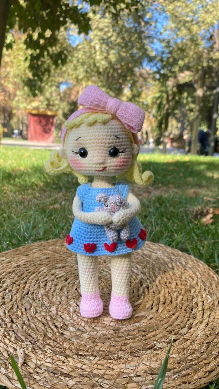 A handmade crocheted doll with blonde hair, a pink bow, blue dress with red hearts, holding a small gray toy.