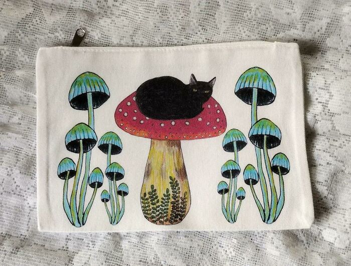 A handmade pouch featuring a black cat resting on a red mushroom surrounded by blue-capped mushrooms.