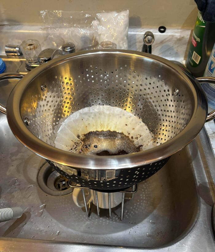 A colander holding a coffee filter over a mug in a sink, a clever solution for making coffee.